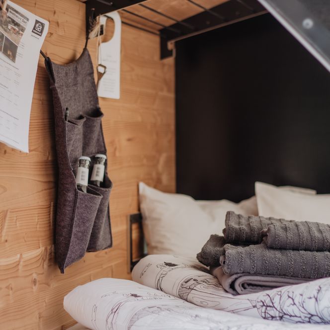 Der Innenraum des "Bett To Go" zeigt ein gemütliches Holzdesign. Es sind ein Bett mit weißem Dekor und eine praktische Hängeaufbewahrung mit Handtüchern und Flaschen zu sehen.