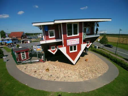 Das Bild zeigt ein auf dem Kopf stehendes, rot-weißes Holzhaus, umgeben von grünem Rasen und einer asphaltierten Straße.