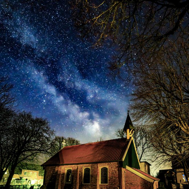 Ein sternklarer Nachthimmel über der alten Backsteinkirche von Spiekeroog, umgeben von Bäumen.